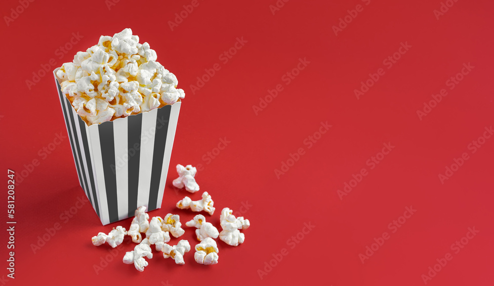 Black white striped carton bucket with tasty cheese popcorn, isolated on red background. Box with scattering of popcorn grains. Fast food, movies, cinema and entertainment concept.