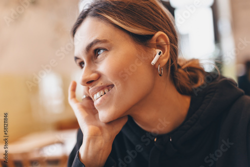 Close up smiling young woman is sitting in a restaurant and putting wireless earphones on. A woman is relaxing during her coffee break. A young woman in restaurant using technologies listening music.