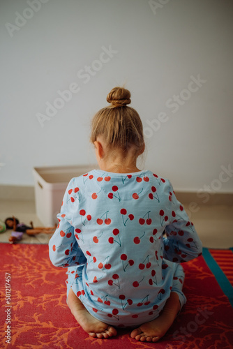 Back view of Cute little girl in blue pyjama with red dots and a hear bun sits on orange carpet and plays with her toys
