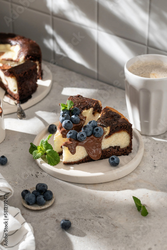 Two pieces of homemade chocolate cheesecake with blueberries, chocolate sauce and mint on a plate with coffee on white kitchen table for morning breakfast in hursh light. Text space, copy space photo