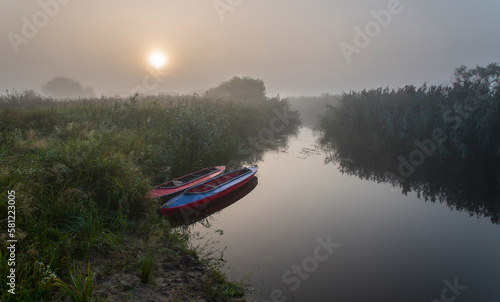 Two multi-colored kayaks at sunrise near the river bank in the morning mist among the river grass.