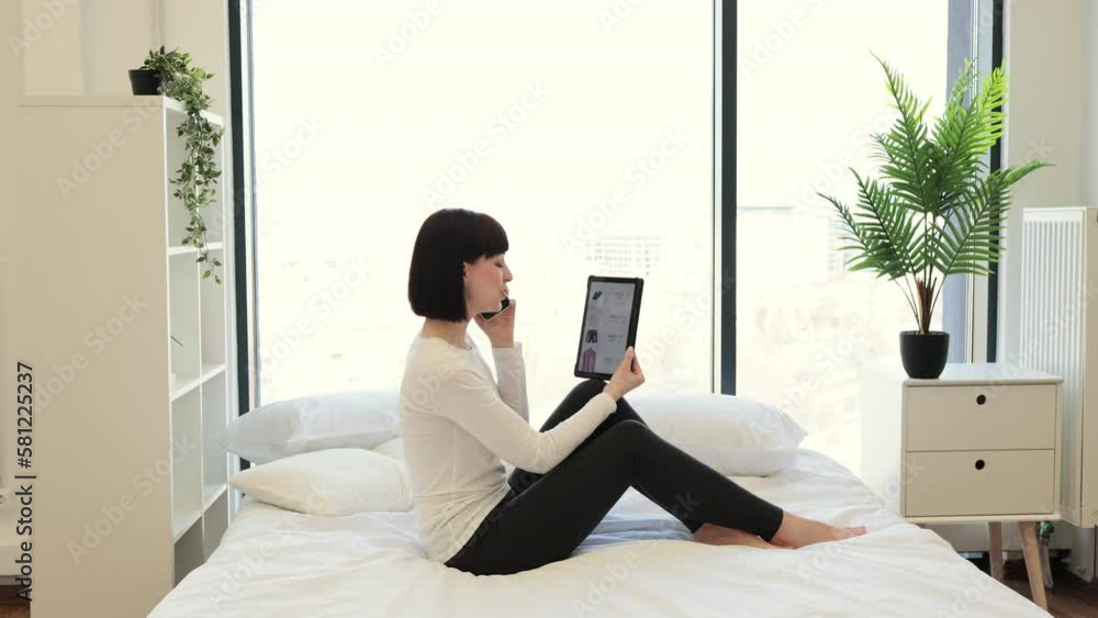 Side view of caucasian woman holding phone while looking at online website with trendy clothes on sale using digital tablet. Casually dressed lady enjoying shopping with big discount at home. Side view of caucasian woman holding phone while looking at online website with trendy clothes on sale using digital tablet. Casually dressed lady enjoying shopping with big discount at home.