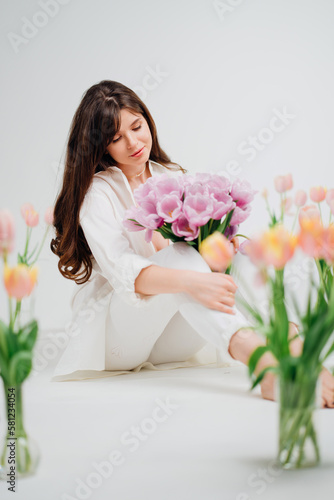 gentle brunette in white sits with a bouquet on the floor with vases of flowers