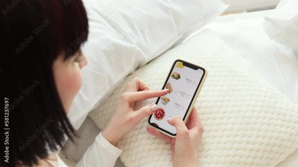 Portrait of beautiful caucasian woman lying on bed and holding modern smartphone in hands. Focus on mobile screen with ecommerce online shop selling food with discount. Portrait of beautiful caucasian woman lying on bed and holding modern smartphone in hands. Focus on mobile screen with ecommerce online shop selling food with discount.