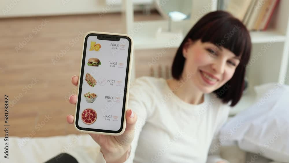 Portrait of beautiful caucasian woman sitting on bed and holding modern smartphone in hands. Focus on mobile screen with ecommerce online shop selling food with discount. Portrait of beautiful caucasian woman sitting on bed and holding modern smartphone in hands. Focus on mobile screen with ecommerce online shop selling food with discount.