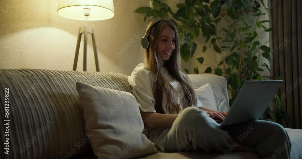 Beautiful middle aged woman sitting on sofa at home, listening to music with wireless headphones and typing on laptop Beautiful middle aged woman sitting on sofa at home, listening to music with wireless headphones and typing on laptop