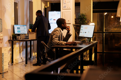 Executive manager analyzing marketing report on computer, checking financial graphs report while working overhours at company strategy. African american businessman planning investment plan photo