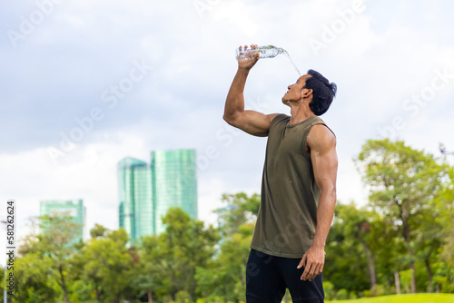Fototapeta Naklejka Na Ścianę i Meble -  Asian man in sportswear drinking and pouring water from a bottle on his face after jogging exercise at public park in the morning. Healthy guy athlete do sport training workout running in the city.