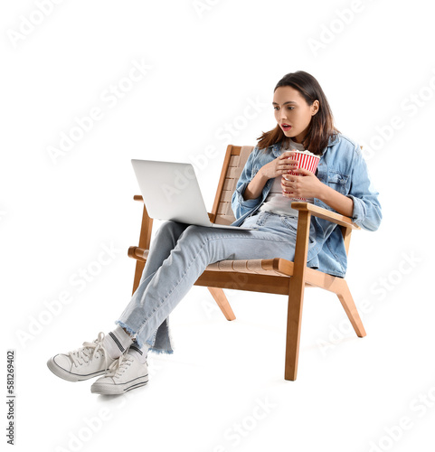 Shocked young woman with popcorn and laptop watching video in armchair on white background