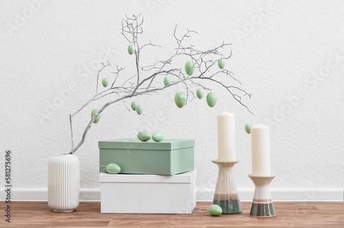 Vase with tree branch, Easter eggs, candles and boxes near light wall
