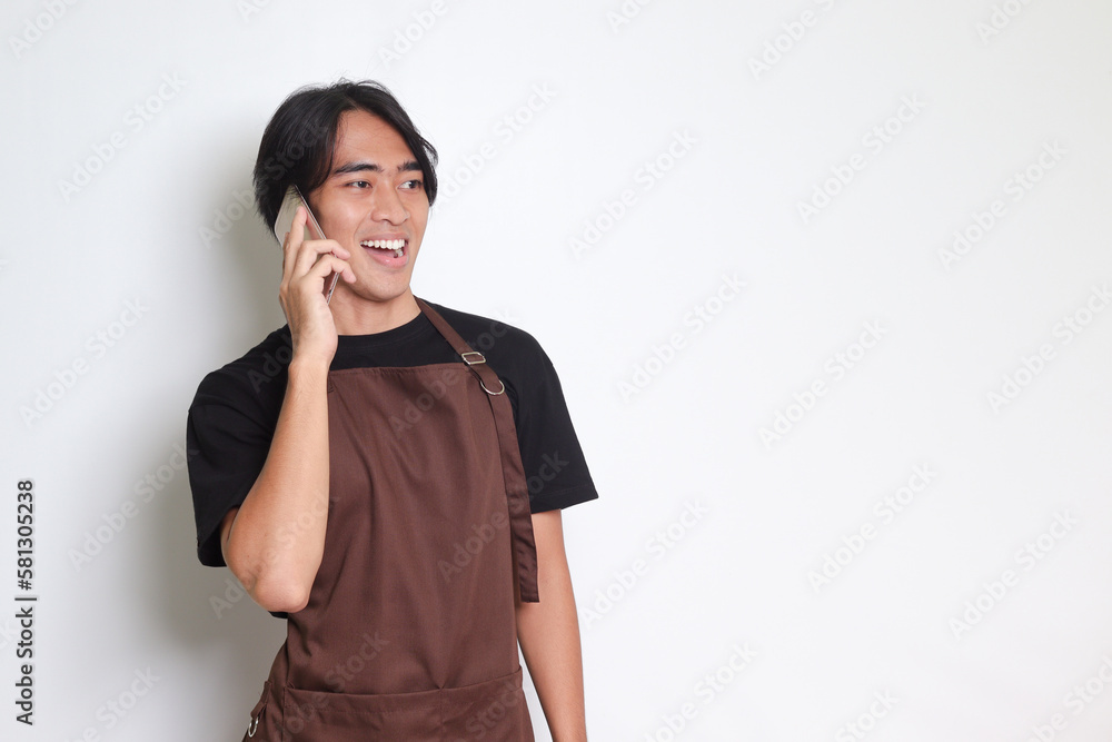 Portrait of attractive Asian barista man in brown apron having conversation while calling someone on mobile phone. Isolated image on white background Portrait of attractive Asian barista man in brown apron having conversation while calling someone on mobile phone. Isolated image on white background