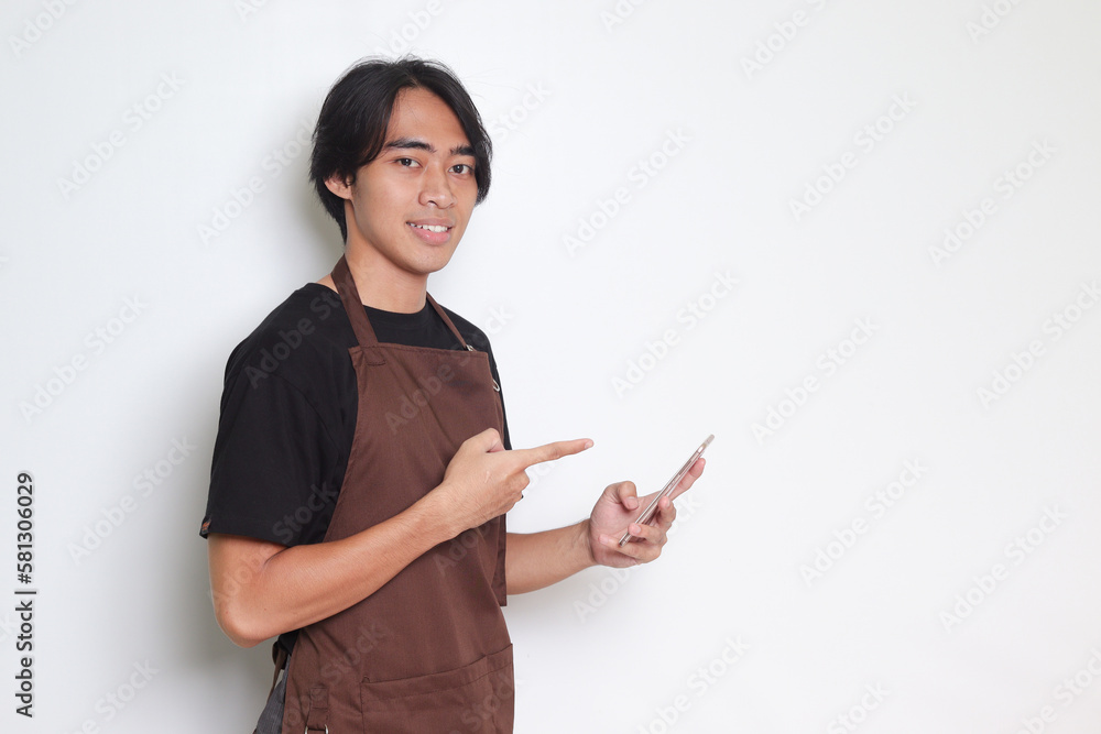 Portrait of attractive Asian barista man in brown apron holding and pointing mobile phone. Isolated image on white background Portrait of attractive Asian barista man in brown apron holding and pointing mobile phone. Isolated image on white background