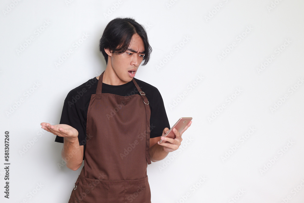 Portrait of shocked Asian barista man in brown apron looking at his mobile phone with surprised expression. Advertising concept. Isolated image on white background Portrait of shocked Asian barista man in brown apron looking at his mobile phone with surprised expression. Advertising concept. Isolated image on white background