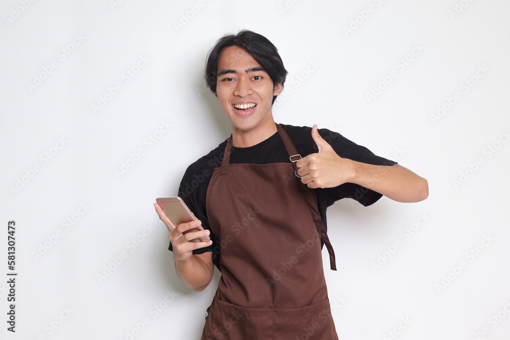 Portrait of attractive Asian barista man in brown apron showing thumb up hand gesture while holding a mobile phone. Advertising concept. Isolated image on white background Portrait of attractive Asian barista man in brown apron showing thumb up hand gesture while holding a mobile phone. Advertising concept. Isolated image on white background