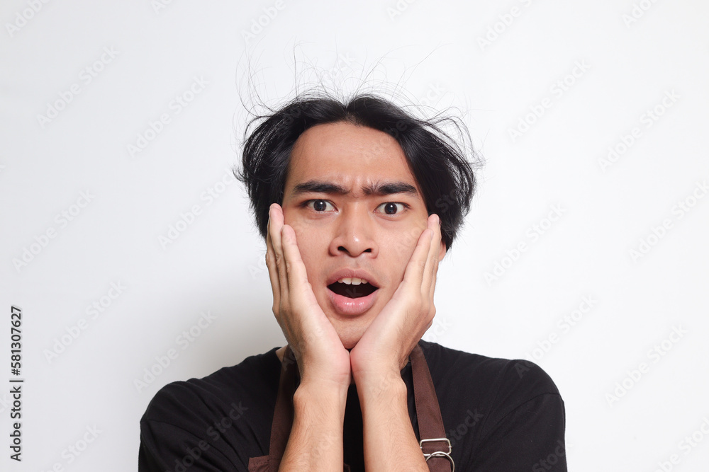 Close up portrait of shocked Asian barista man in brown apron holding hands on cheeks, feeling surprised with open mouth. Isolated image on white background