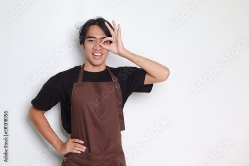 Portrait of attractive Asian barista man in brown apron showing ok hand gesture and smiling looking at camera. Advertising concept. Isolated image on white background