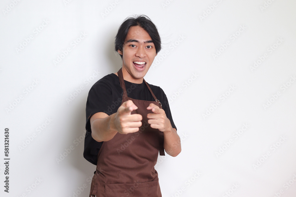 Portrait of attractive Asian barista man in brown apron pointing forward to the camera with finger. Advertising concept. Isolated image on white background Portrait of attractive Asian barista man in brown apron pointing forward to the camera with finger. Advertising concept. Isolated image on white background