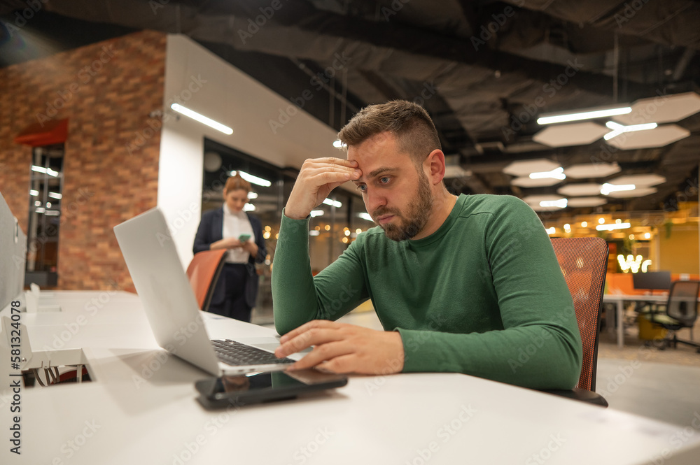 Bearded man holding his head while working on a laptop in an open space office. Red-haired caucasian woman uses smartphone. Bearded man holding his head while working on a laptop in an open space office. Red-haired caucasian woman uses smartphone.