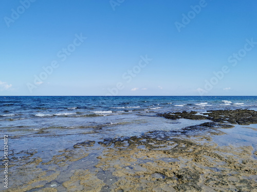 The rocky coast of the Mediterranean Sea from long-hardened lava, waves, clear water against a blue sky with clouds.