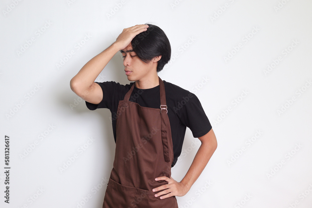 Portrait of attractive Asian barista man in brown apron holding hand on forehead, forgets something, and regrets himself. Isolated image on white background Portrait of attractive Asian barista man in brown apron holding hand on forehead, forgets something, and regrets himself. Isolated image on white background
