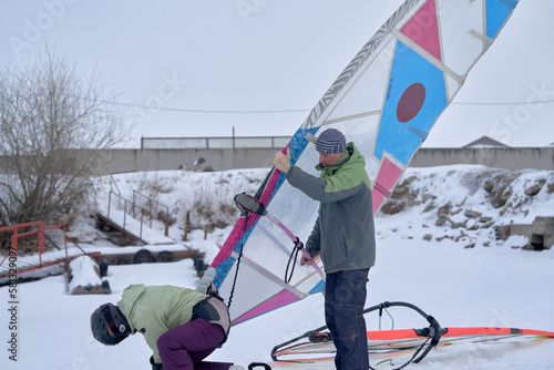 A middle-aged man and woman, snowsurfers, prepare sailboards for skiing in the snow. Preparing to ride a sailboard in the snow on a cloudy winter day.