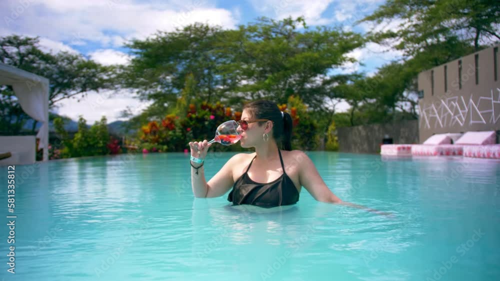 Young woman in sunglasses drinks cocktail in pool, push-in portrait Young woman in sunglasses drinks cocktail in pool, push-in portrait