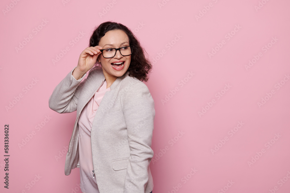 Charming dark-haired middle-aged woman smiling a toothy smile, looking aside, wearing stylish trendy eyeglasses and elegant light gray suit, isolated on pink background with free advertising space