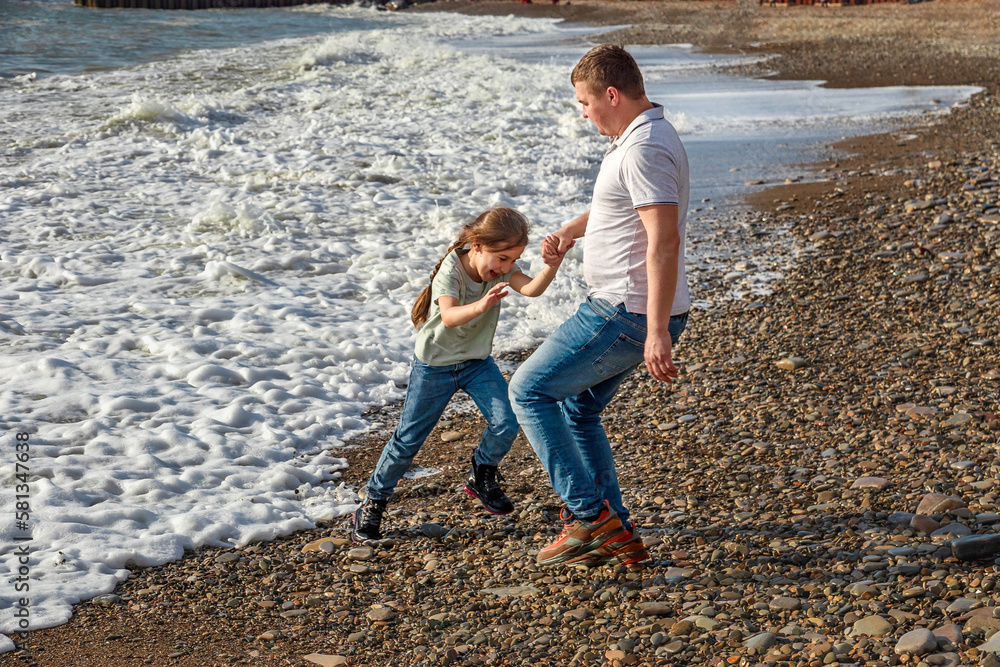 Father and daughter play on the beach in the afternoon. Father and daughter are running on the waves of the surf. Dad and daughter lead a carefree, happy, cheerful, smiling lifestyle.