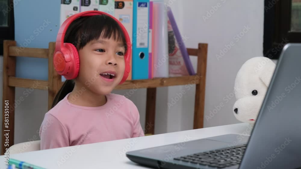 Cute elementary school girl wearing headphones and using a laptop computer. Happy Asian kids study online interactively with laptop computer or homeschooling, listening to music or playing games. Cute elementary school girl wearing headphones and using a laptop computer. Happy Asian kids study online interactively with laptop computer or homeschooling, listening to music or playing games.