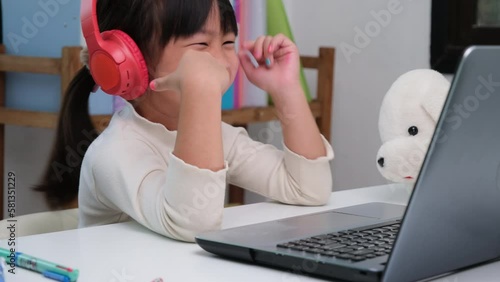 Cute elementary school girl wearing headphones and using a laptop computer. Happy Asian kids study online interactively with laptop computer or homeschooling, listening to music or playing games.
