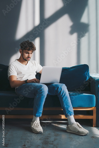 Young man looking at the screen of a laptop computer while relaxing on a comfortable sofa at home in casual wears. Modern lifestyle. Modern communication