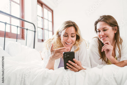 Happy two friends Caucasian young woman in white bathrobes using a smartphone together with fun and smiles while lying on the bed at home. LGBTQ couples leisure spa and treatment activities.