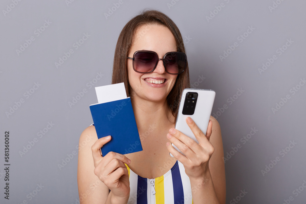 Portrait of happy cheerful young adult woman wearing swimsuit and sunglasses holding passport isolated on gray background, using mobile phone, booking voucher for abroad country Portrait of happy cheerful young adult woman wearing swimsuit and sunglasses holding passport isolated on gray background, using mobile phone, booking voucher for abroad country