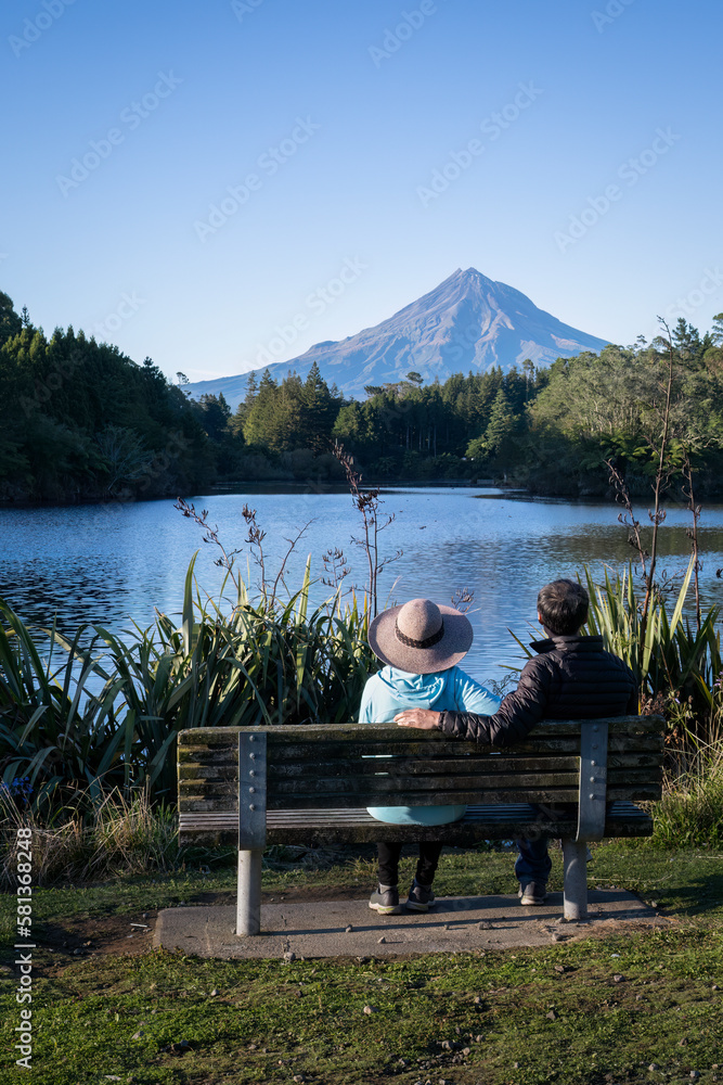 Couple sitting on the bench and enjoying the view of Mt Taranaki at Lake Mangamahoe. New Plymouth. Vertical format. Couple sitting on the bench and enjoying the view of Mt Taranaki at Lake Mangamahoe. New Plymouth. Vertical format.