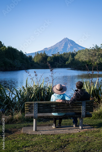 Couple sitting on the bench and enjoying the view of Mt Taranaki at Lake Mangamahoe. New Plymouth. Vertical format.