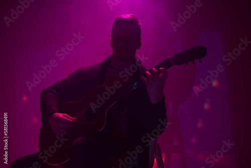 Musician playing acoustic guitar in a foggy club with colorful lights.