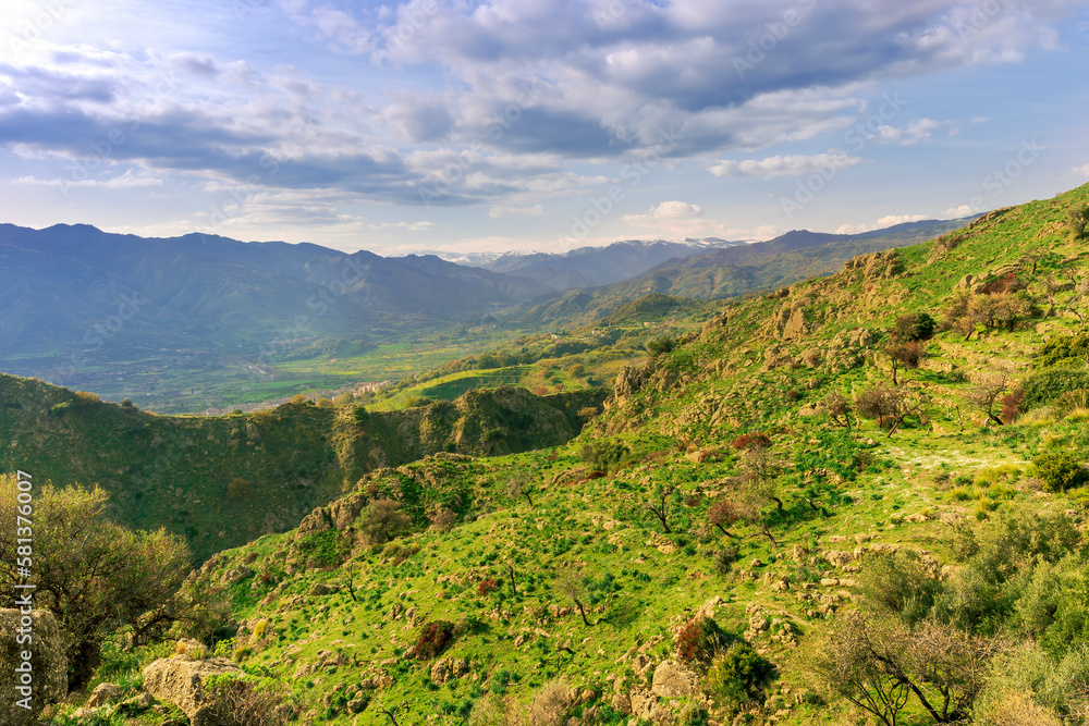 picturesque view in a green mountain valley among slopes and hills with sunny plantations on a rocky plato and nice cloudy sunset on background