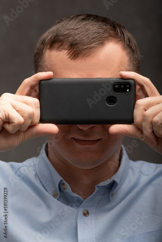 Young man on gray background holding mobile smartphone on front of eyes. Portrait of man with phone near his face