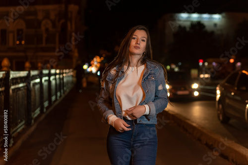Stylish attractive young woman in denim jacket, evening city background.Portrait of girl student walking on night street