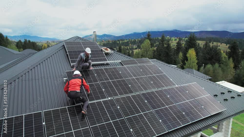 Technicians building photovoltaic solar module station on roof of house. Men electricians in helmets installing solar panel system outdoors. Concept of alternative and renewable energy. Aerial view.