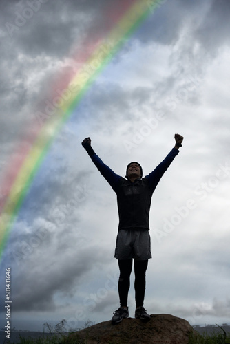 Triumph at the top. A young man celebrating at the top of a mountain under a rainbow.