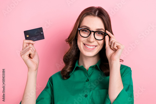 Photo of toothy beaming woman with wavy hairdo dressed green shirt touching eyewear holding plastic card isolated on pink background