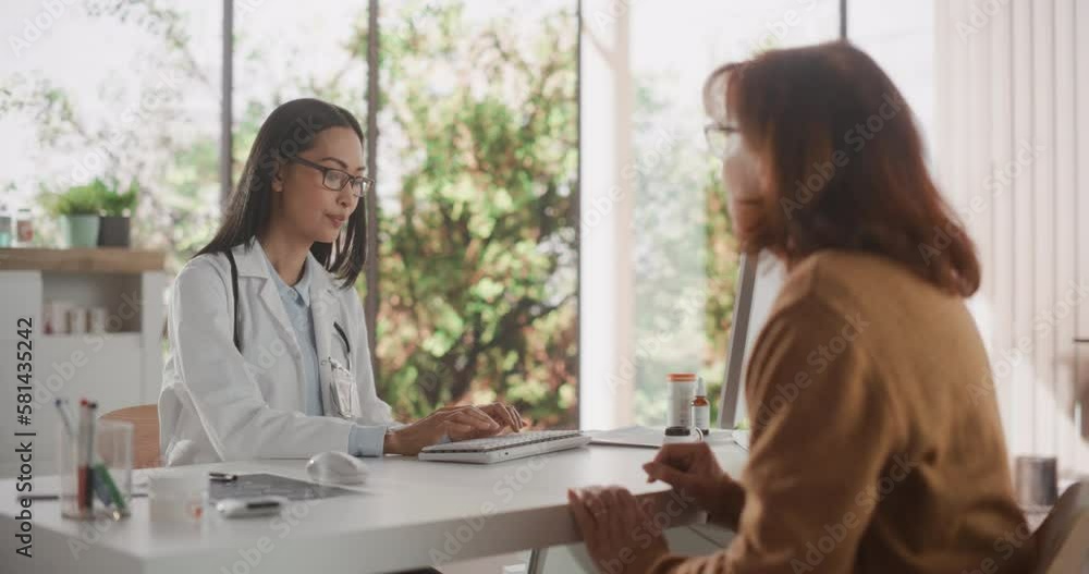 Young Asian Physician is Prescribing Medicine for an Adult Female Patient and Listening to Her Feedback During Consultation in a Health Clinic. Doctor in Lab Coat Sitting Behind a Desk in Hospital Young Asian Physician is Prescribing Medicine for an Adult Female Patient and Listening to Her Feedback During Consultation in a Health Clinic. Doctor in Lab Coat Sitting Behind a Desk in Hospital