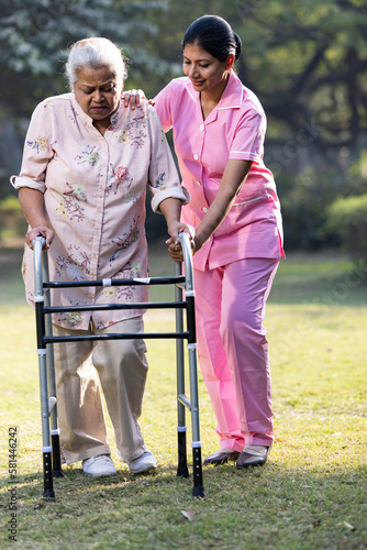 Female nurse helping senior woman with mobility walker at nursing home garden. © G-images