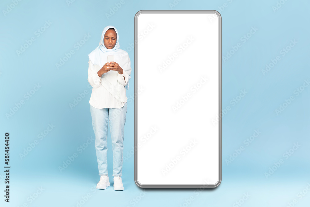 Black muslim woman using cellphone while standing near big white smartphone screen over blue studio wall, full length Black muslim woman using cellphone while standing near big white smartphone screen over blue studio wall, full length