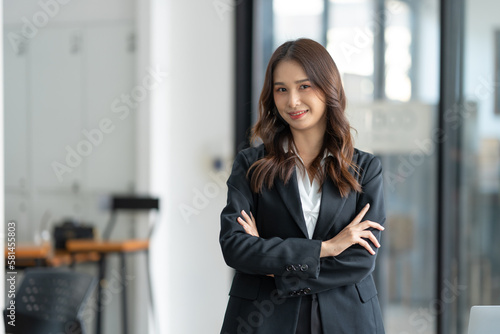 Asian businesswoman or freelancer standing near the desk in the office Dressed in formal clothes, crossed arms, looking at the camera, smiling friendly