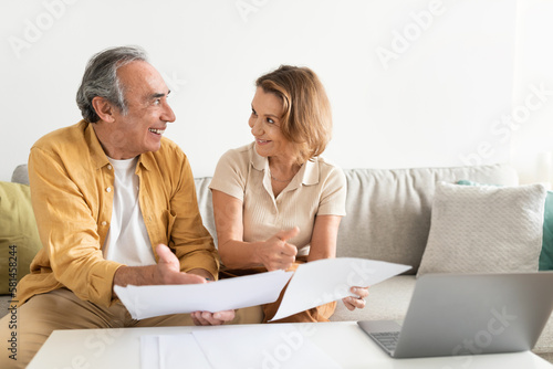 Senior couple holding documents reading paper bills, paying bank loan online, using laptop at home, free space
