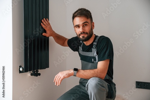 Looking at the camera. Plumber worker installing heating radiator in empty room of a newly built house