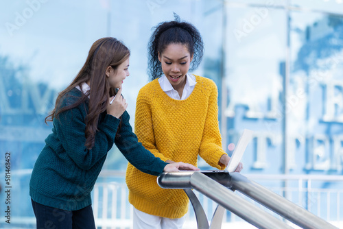 Two women standing talking in front of outdoor office with tablet computer Young woman discussing work with friends outside office