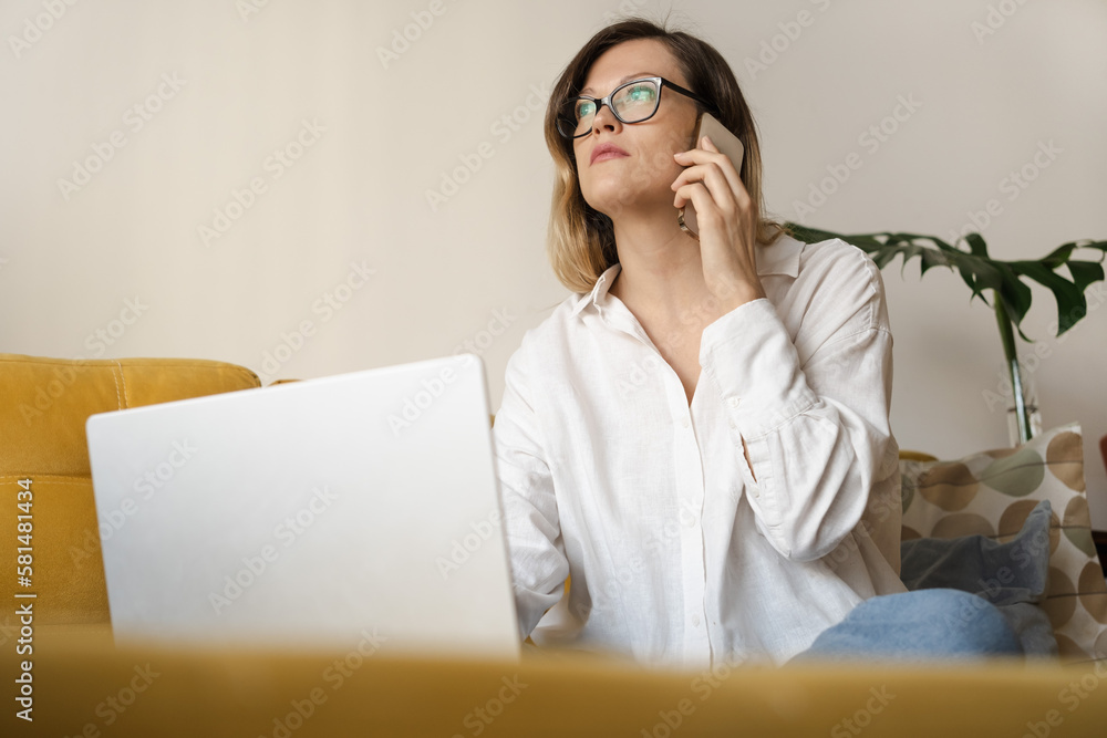 Young freelancer woman wearing eyeglasses sitting on yellow sofa indoors working with laptop and cellphone, thinking out. Work at home office, in distance concept. Copy space. Young freelancer woman wearing eyeglasses sitting on yellow sofa indoors working with laptop and cellphone, thinking out. Work at home office, in distance concept. Copy space.